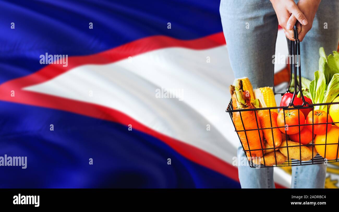 Woman is holding supermarket basket, American Samoa waving flag background. Economy concept for fresh fruits and vegetables. Stock Photo