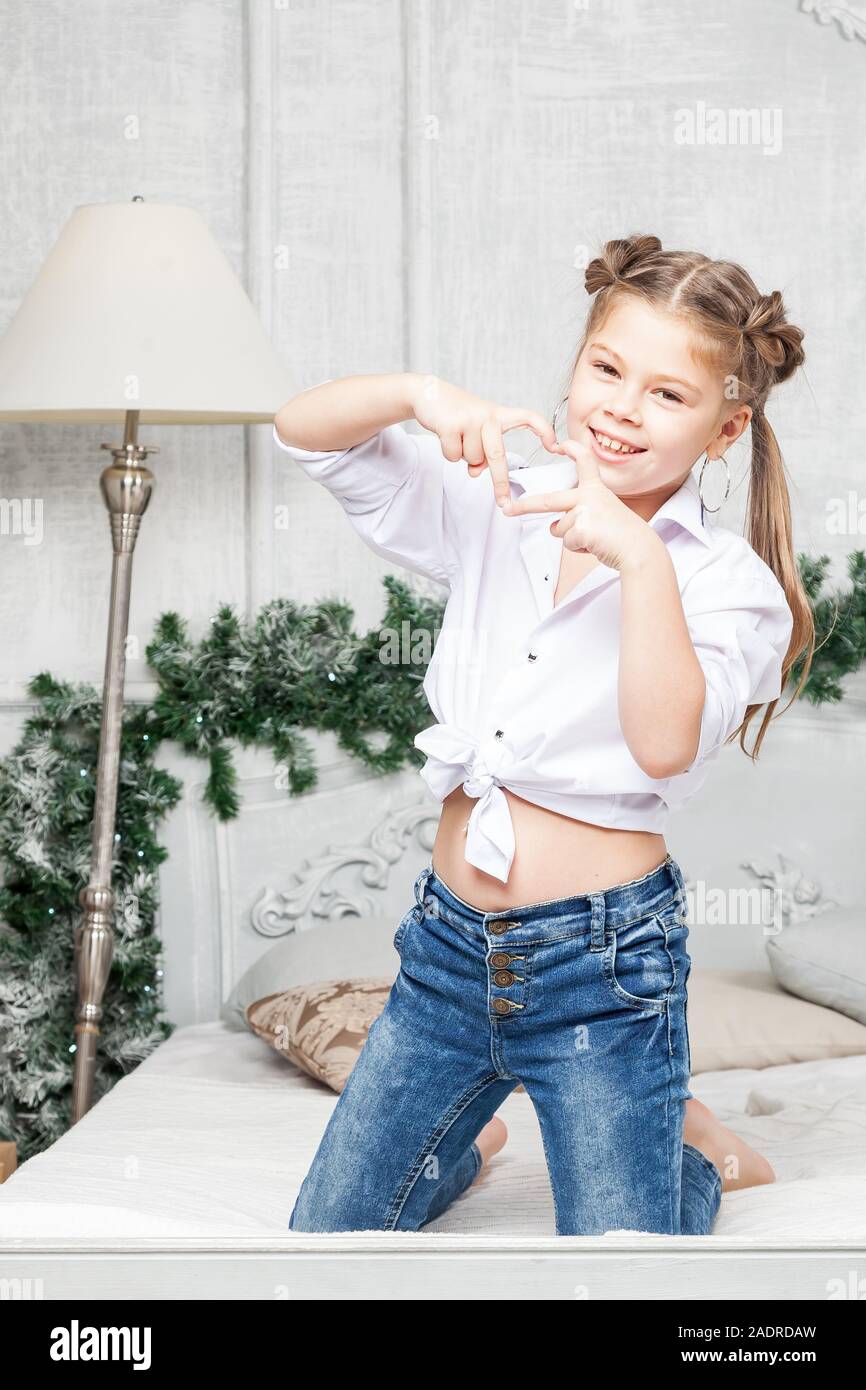 Little cute beautiful girl having fun and kneeling on the bed in blue jeans  and a white shirt, showing heart and love with her fingers in the room wit  Stock Photo -