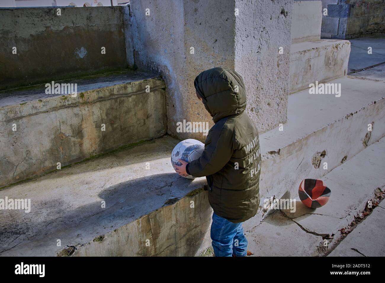 Boy in blue trousers and green jacket playing ball on cement stairs. Stock Photo