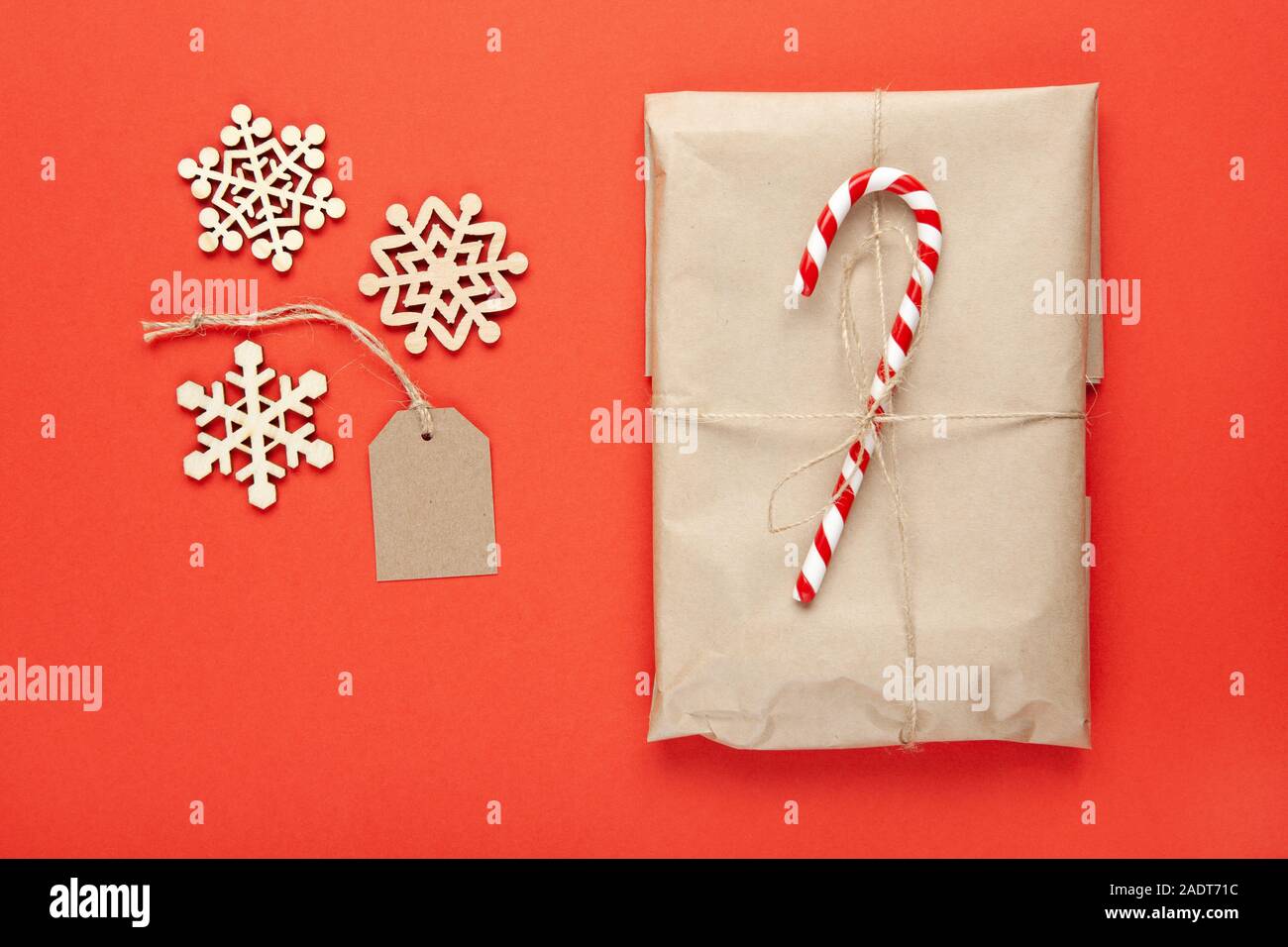 Christmas gift in craft paper, decorated with candy cane, wooden snowflakes and craft tag on bright red background. Festive, New Year, sales concept. Stock Photo