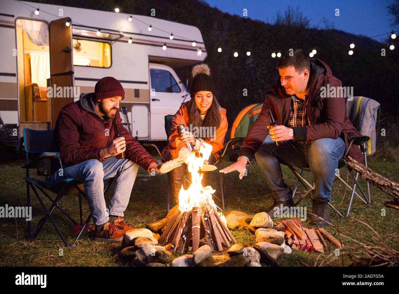 Group of friends enjoying the fresh air of the mountains around camp fire.  Retro camper van with light bulbs Stock Photo - Alamy