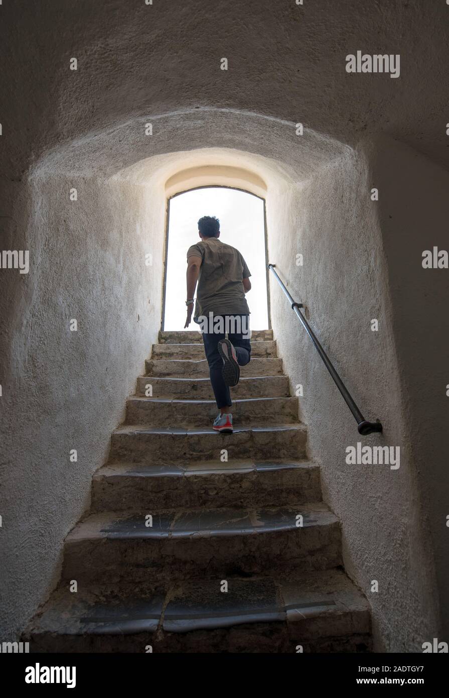 Man running away up a steep flight of stone stairs towards daylight through an arched exit at the top viewed from below Stock Photo