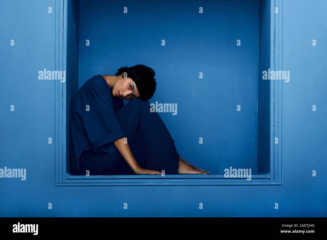 Fashion shot of young beautiful woman sitting in niche on blue background. Studio shot. Stock Photo