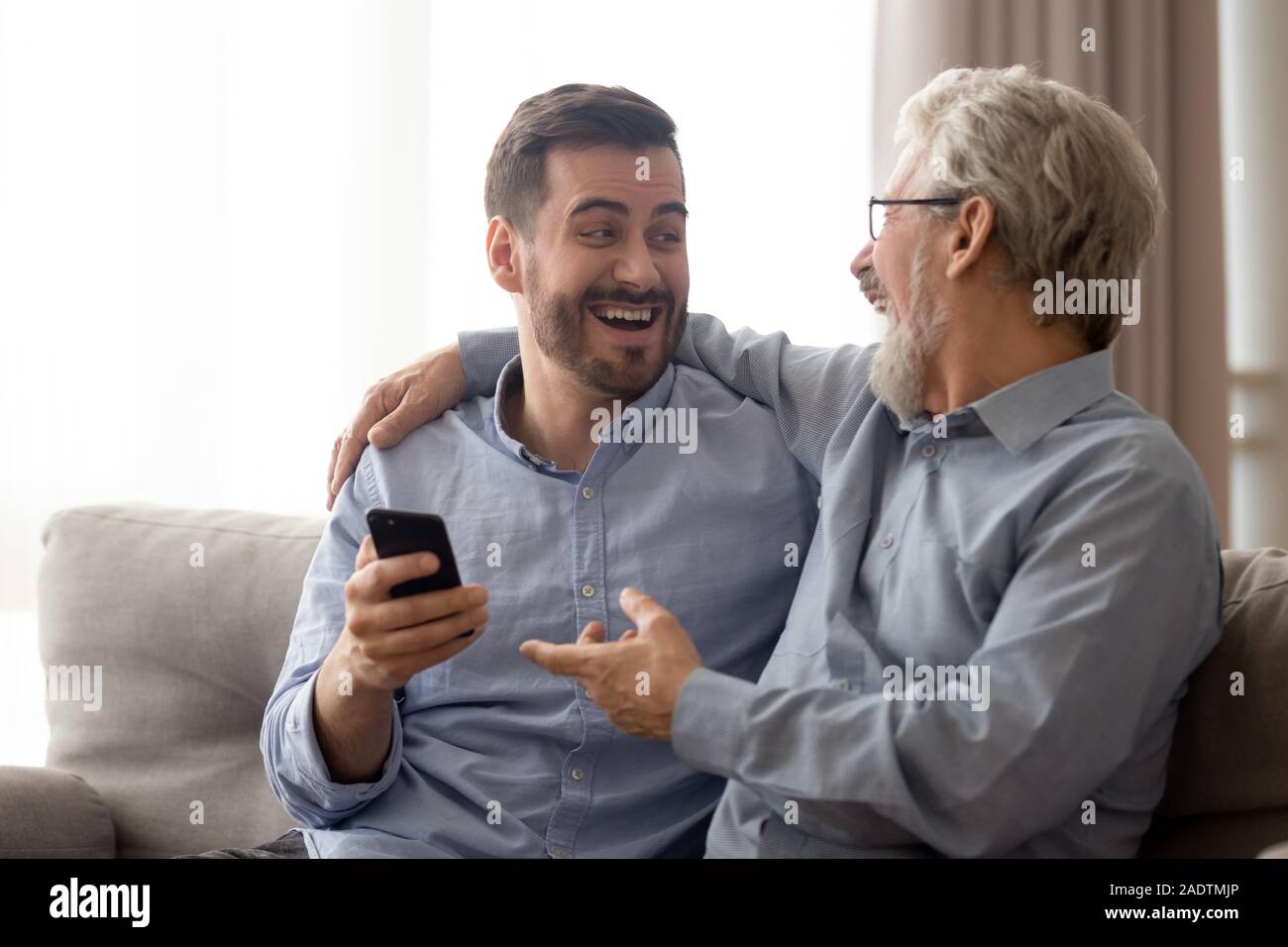 Different generations relatives men using smartphone laughing sit on couch Stock Photo