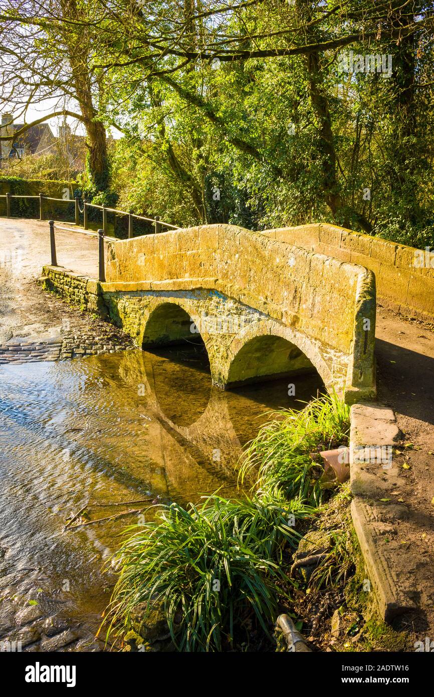 The old stone foot-bridge spans the slowly flowing ByBrook stream which forms part of a ford for vehicular crossings in the quiet village of Lacock i Stock Photo