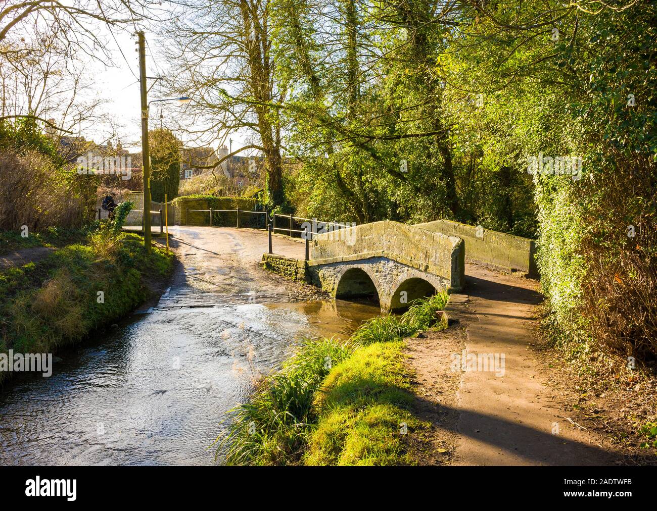 The old stone foot-bridge spans the slowly flowing ByBrook stream which forms part of a ford for vehicular crossings in the quiet village of Lacock i Stock Photo