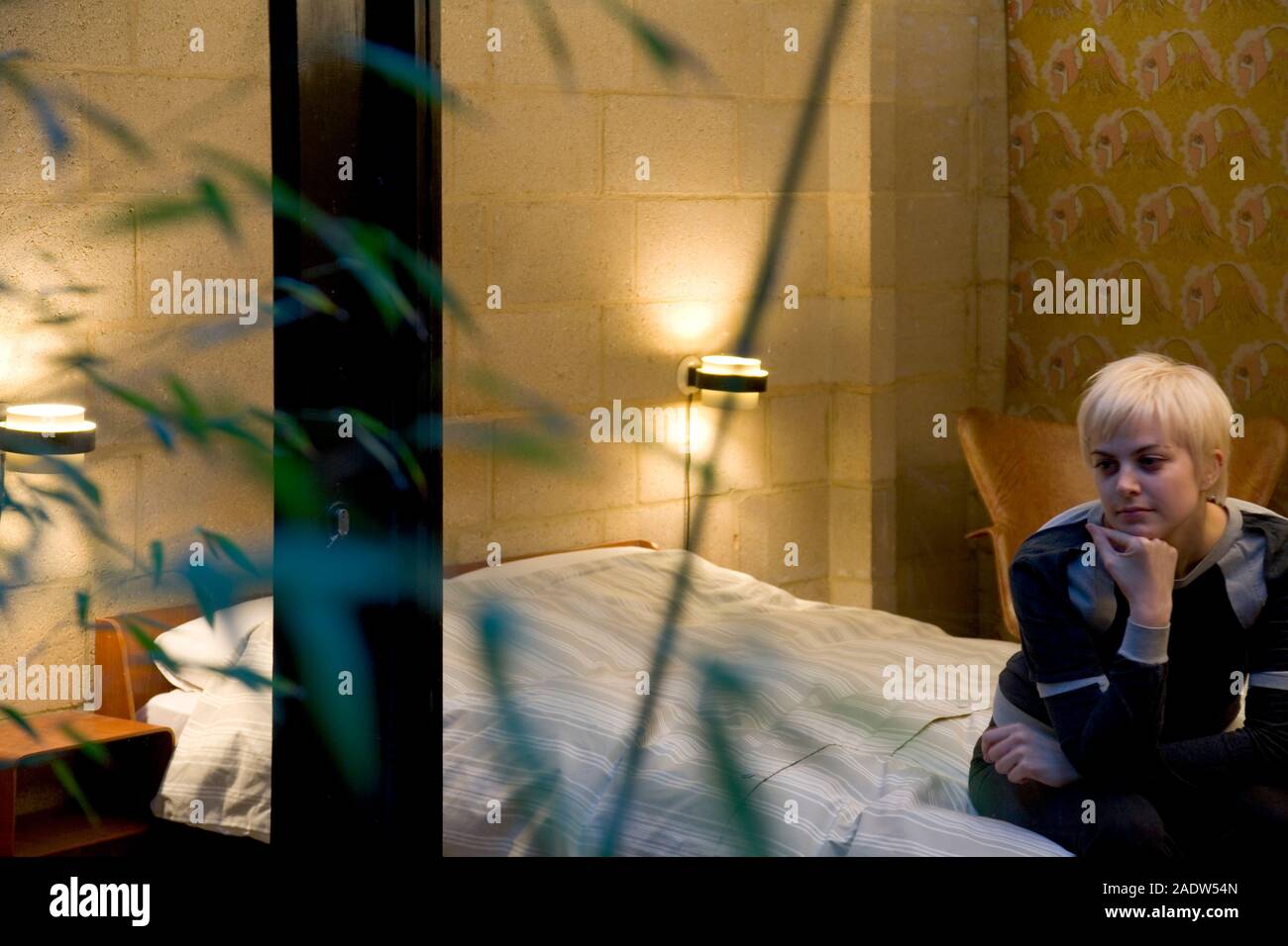 A young woman, 20's, sitting alone on a bed seen through a window and foliage Stock Photo