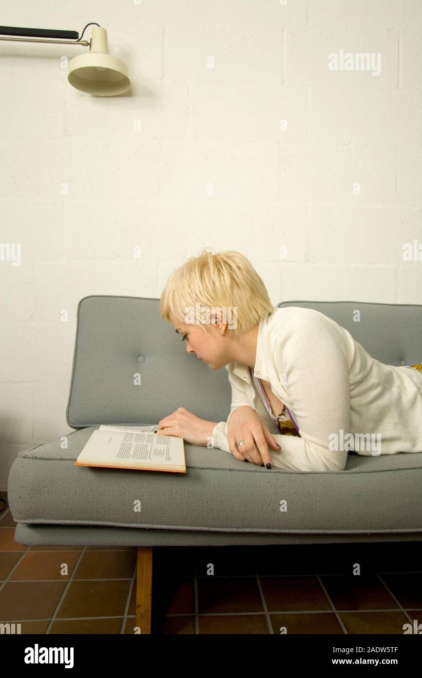 Stylish young woman, 20's, in a simple 1960's retro style domestic interior, reading a book, lying on a sofa Stock Photo