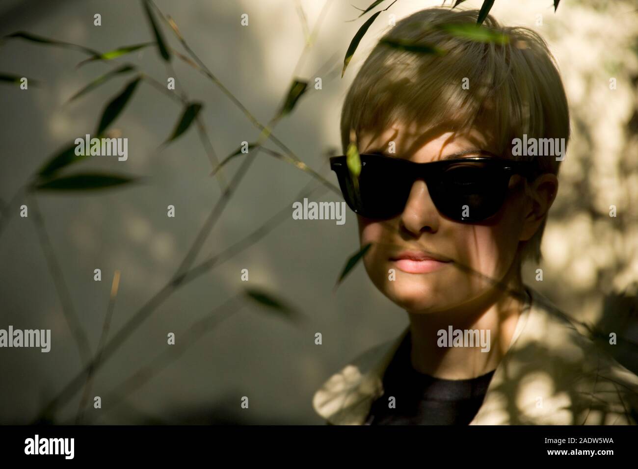 Stylish young woman, 20's, wearing 1950's style trenchcoat and sun glasses, seen through foliage Stock Photo