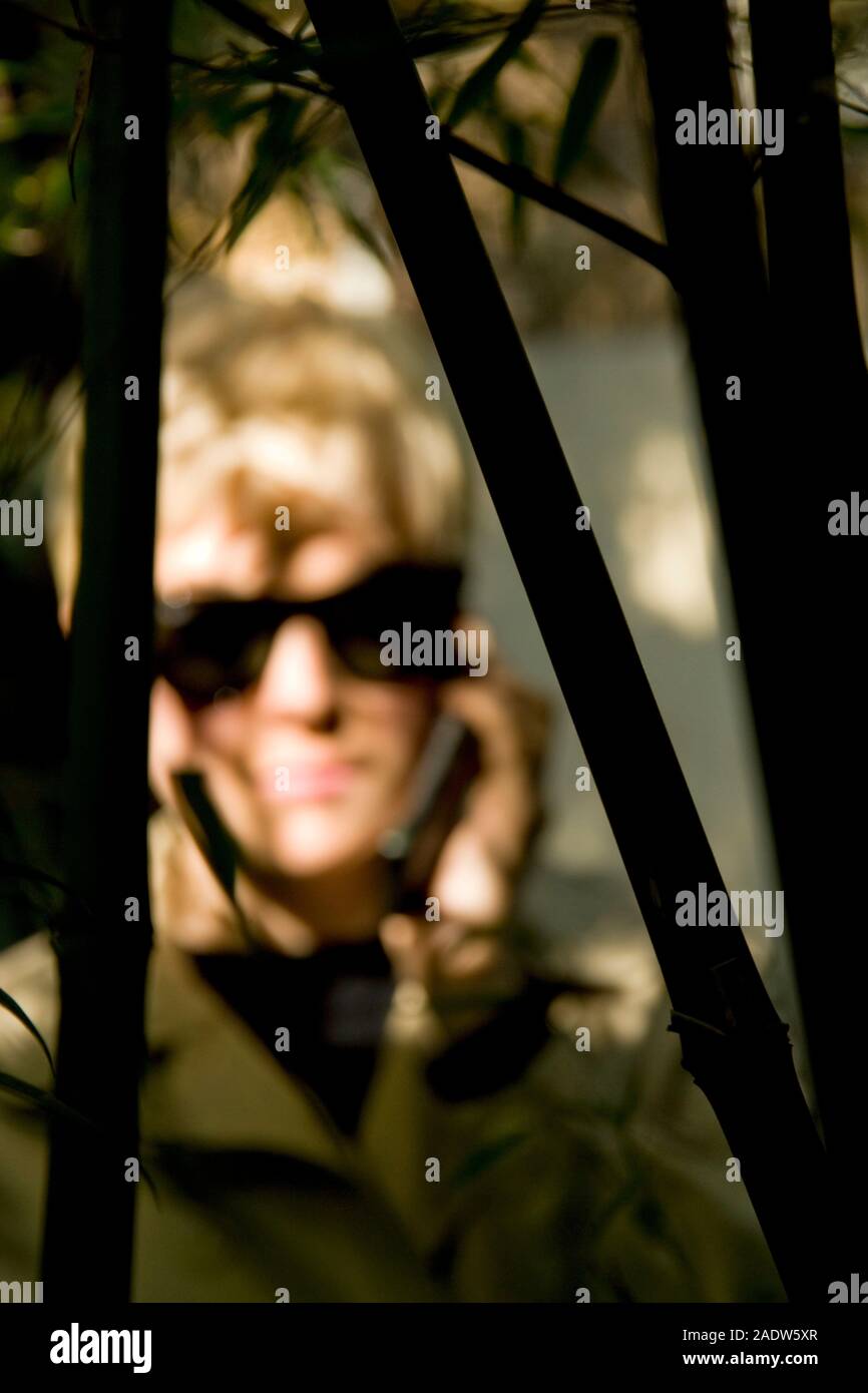 Stylish young woman, 20's, wearing 1950's style trenchcoat and sun glasses, seen through foliage using mobile telephone Stock Photo