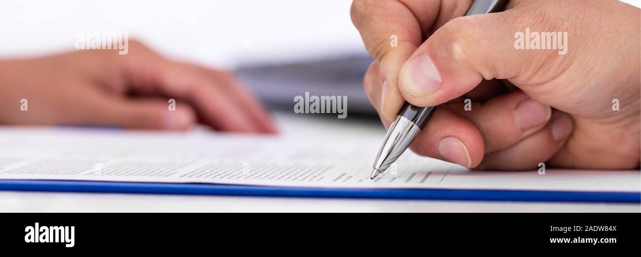 panorama of a man, signing a document on a blue clipboard, hand of a woman in the background Stock Photo