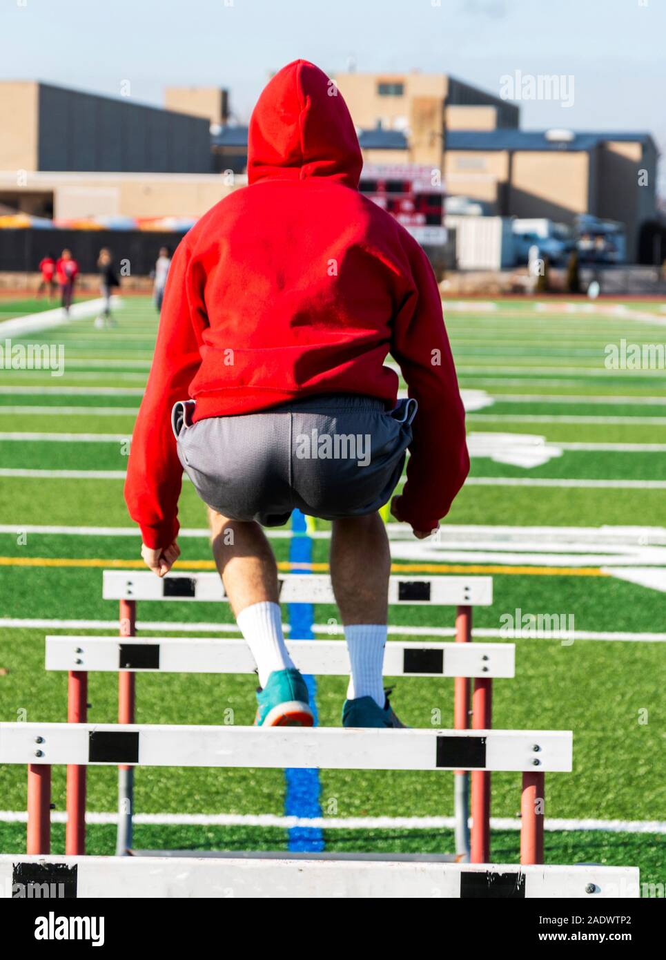 An athlete wearing a red hooded sweatshirt is jumping over hurdles on a green turf field with the view from behind him. Stock Photo