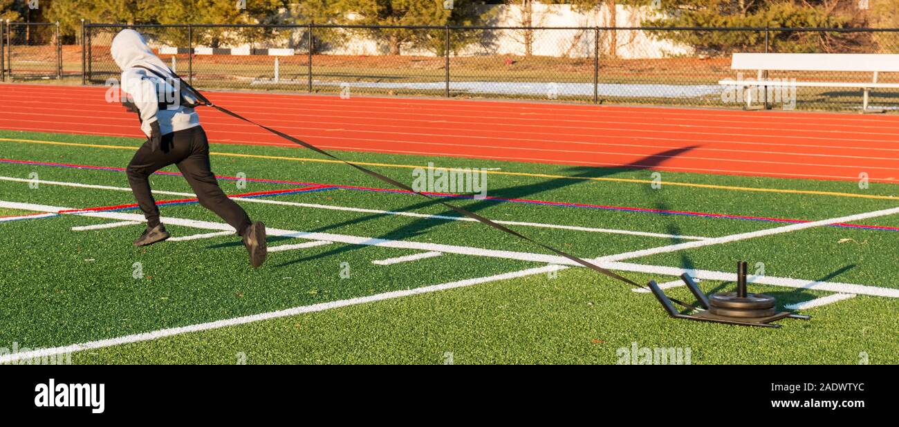 A high school track and field athlete is pulling a sled with weights on it across a green turf field for strength and speed work during practice. Stock Photo