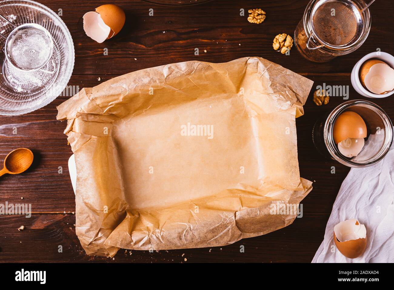 Unfilled baking dish covered with parchment on rustic dark wooden background among ingredients and utensils for making pastry. Stock Photo