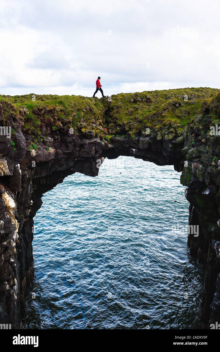 Tourist walks over a natural rock bridge connecting basalt cliffs in Arnarstapi, Iceland, with Atlantic Ocean in the background. Stock Photo