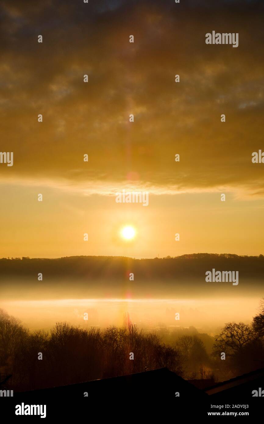 A beautiful, dramatic winter sunrise across a mist filled valley. The rising sun is captured between a distant hill and low cloud above the valley fog Stock Photo