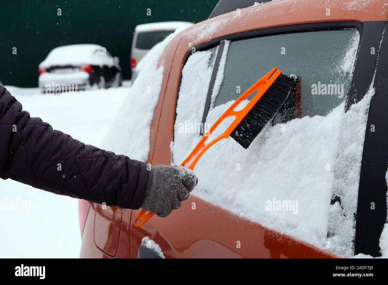 Brush in mans hand. Man in gray gloves clears orange car from snow. Remove lot of snow from  auto. Stock Photo