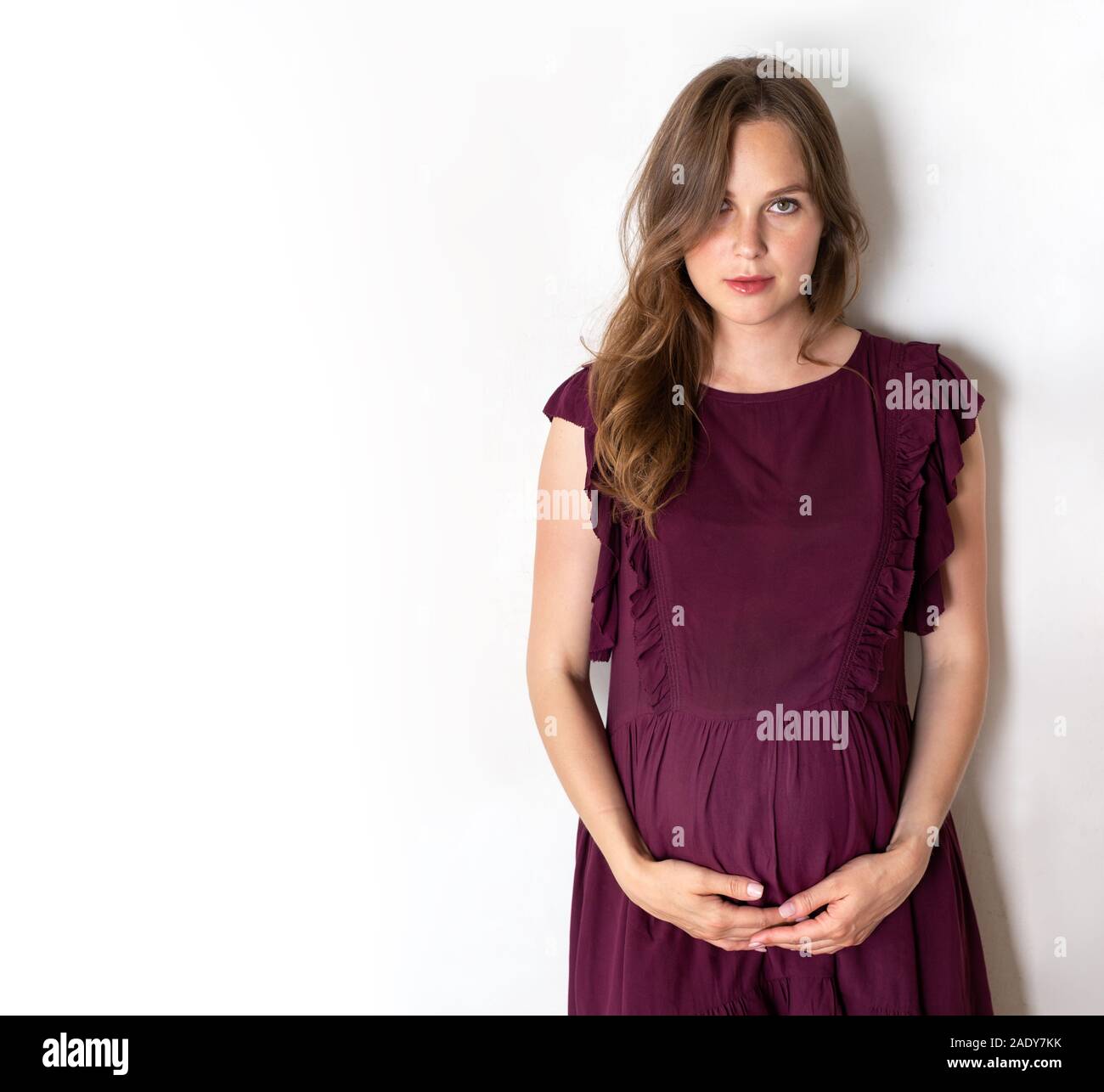 a pregnant girl stands against a white wall. hands holds its big stomach Stock Photo