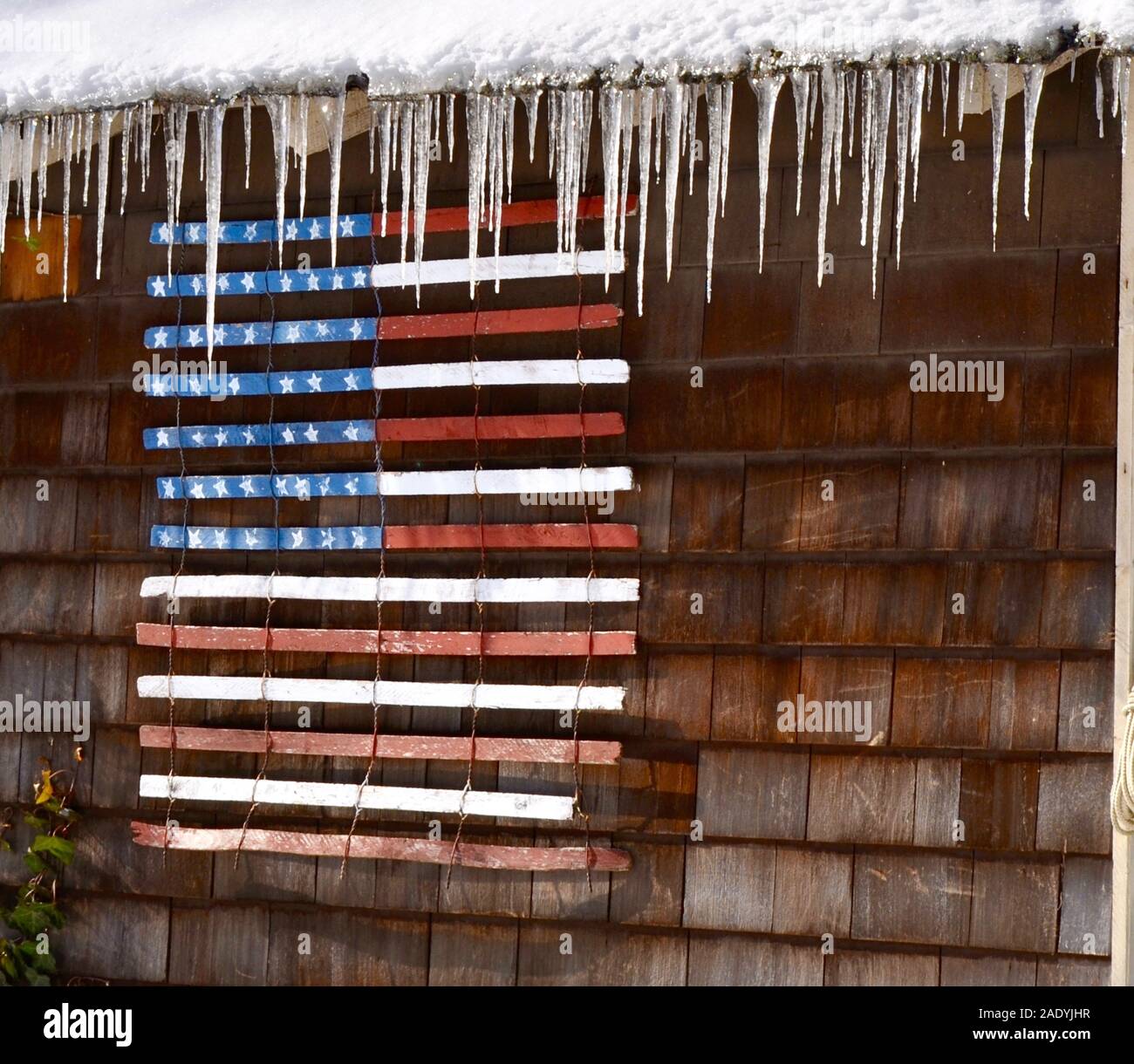 American flag painted on a snow fence hangs from a shed eave trimmed with icicles. Stock Photo