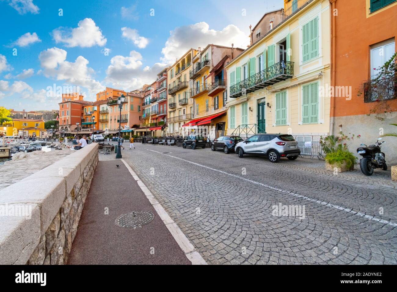 Tourists walk a cobbled street in front of a row of colorful apartments and cafes in Villefranche Sur Mer, on the Mediterranean Coast of France Stock Photo