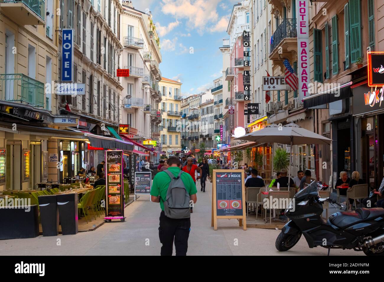 French citizens and tourists walk through a colorful, narrow alley of shops, hotels and cafes as the lights come on at dusk in Nice, France. Stock Photo