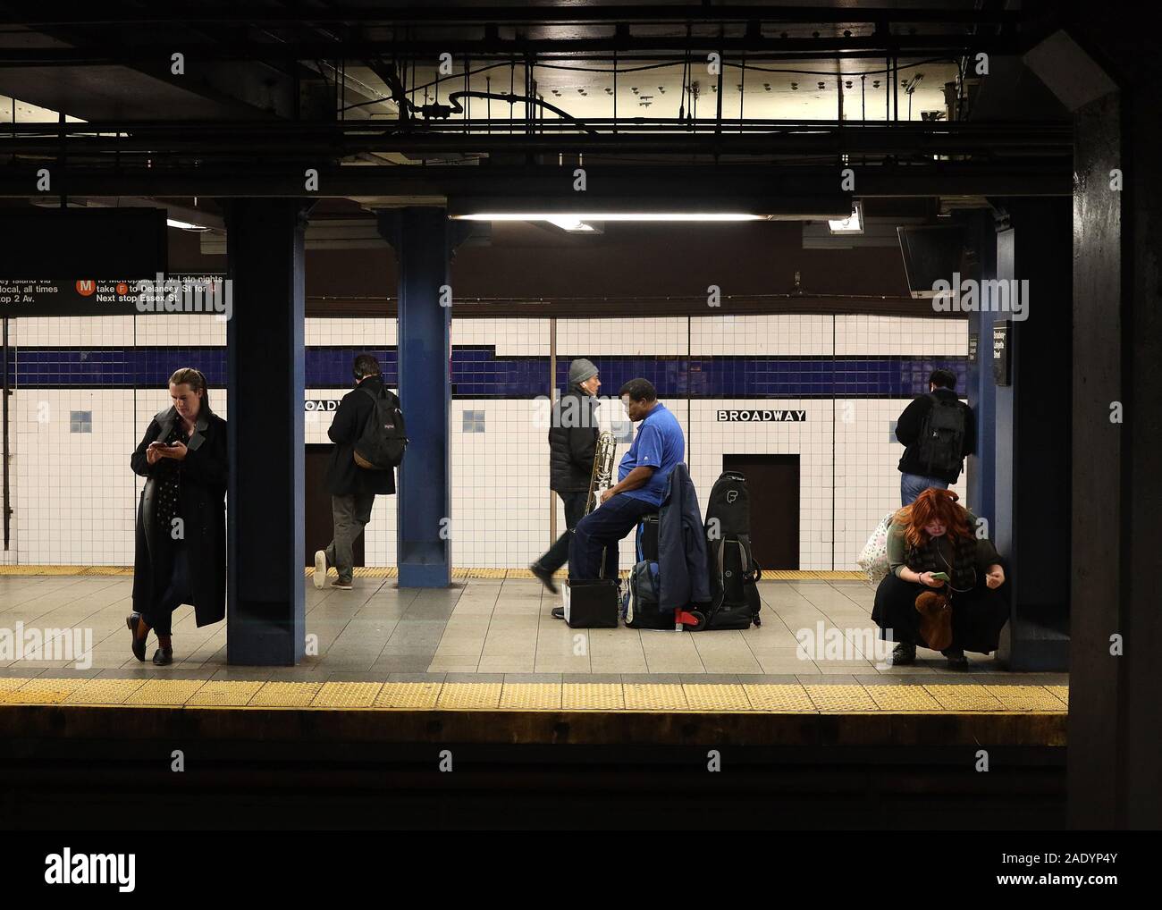 New York City, NY - Novermber 26, 2019: Unidentified trumpetist sitting on his luggage while other unidentified people look at their phones in Manhatt Stock Photo