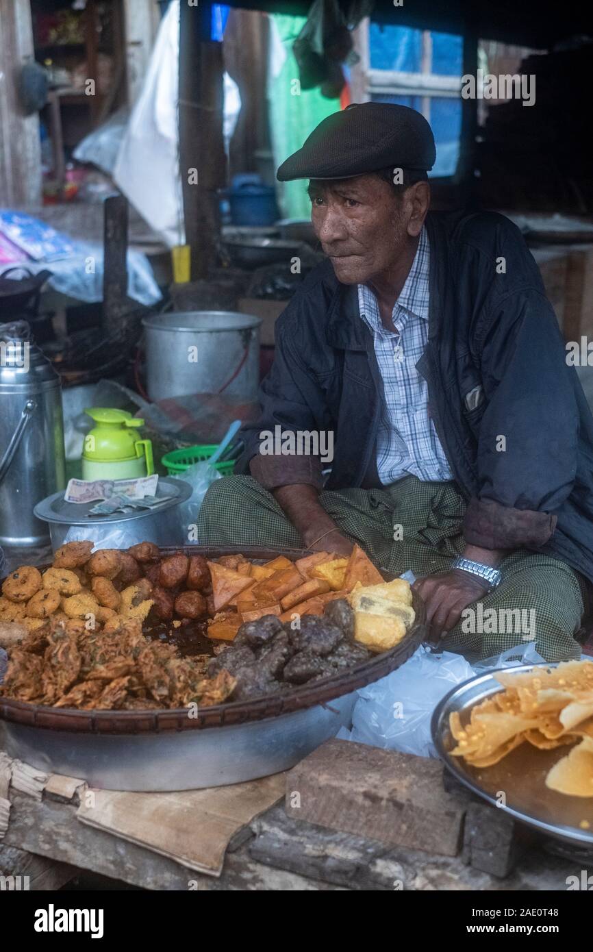 An older Burmese man in a cap sells various fried foods from a stall in a village market in Kanne, northwestern Myanmar (Burma) by the Chindwin River Stock Photo