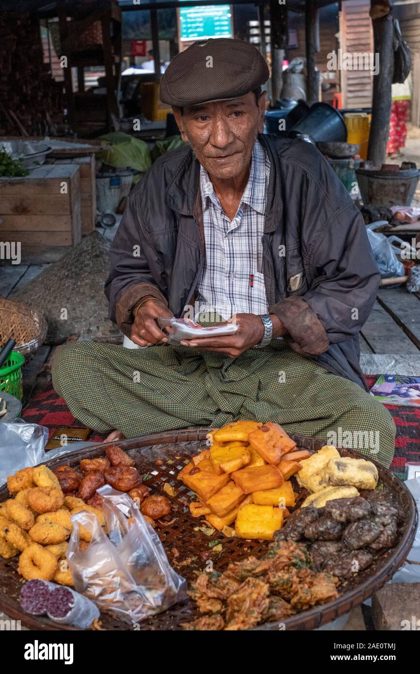 An older Burmese man in a cap sells various fried foods from a stall in a village market in Kanne, northwestern Myanmar (Burma) by the Chindwin River Stock Photo