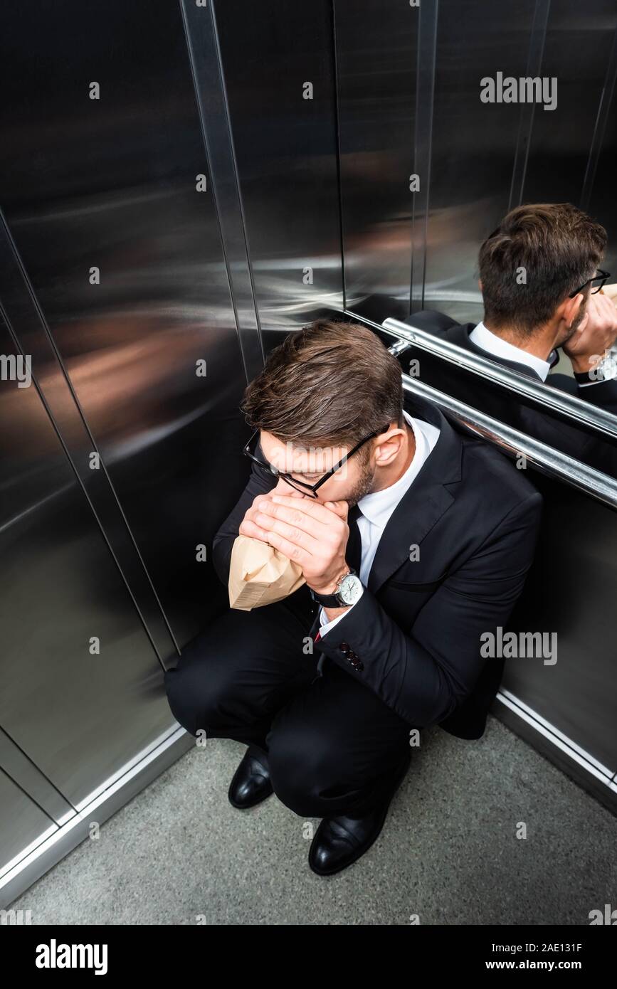 high angle view of scared businessman in suit with claustrophobia breathing in paper bag in elevator Stock Photo