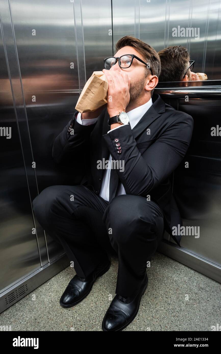 scared businessman in suit with claustrophobia breathing in paper bag in elevator Stock Photo