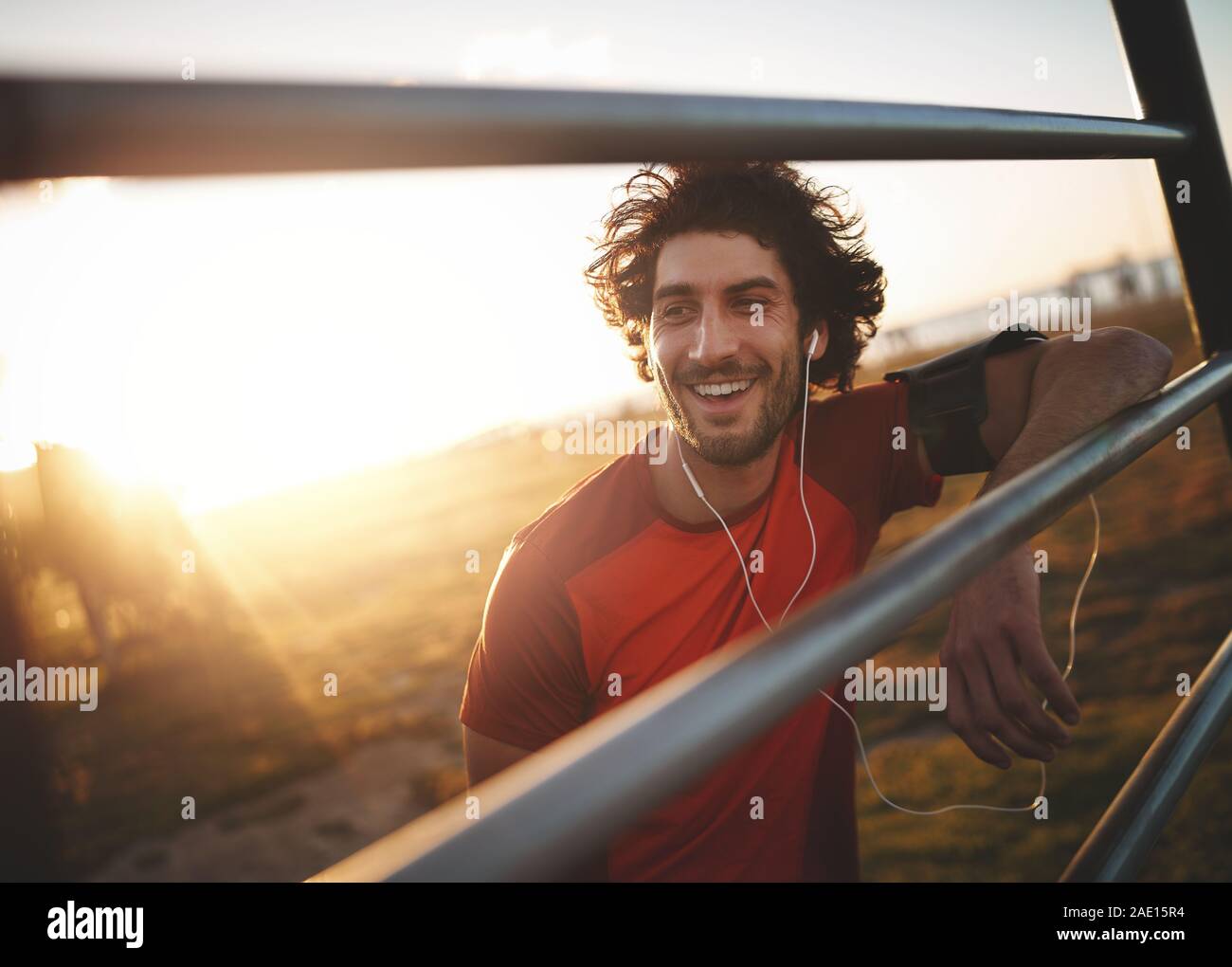 Portrait of a smiling young male athlete with earphones in his ears leaning on horizontal bars resting after run Stock Photo