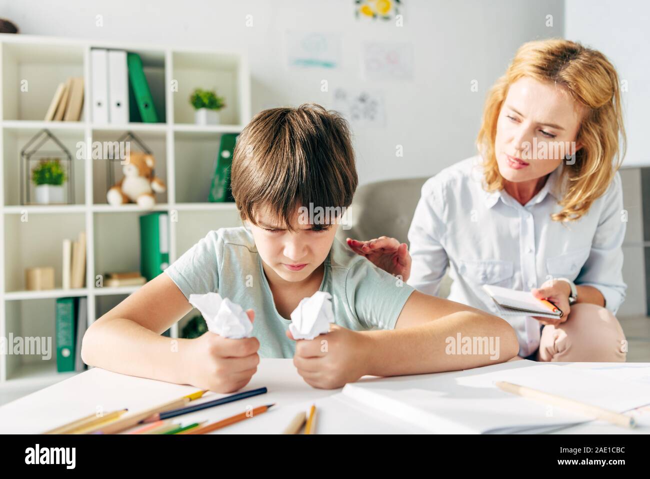irritated kid with dyslexia holding crumpled papers and child psychologist talking to him Stock Photo