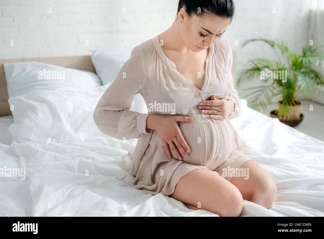 beautiful worried pregnant woman in nightie touching belly while sitting on bed Stock Photo