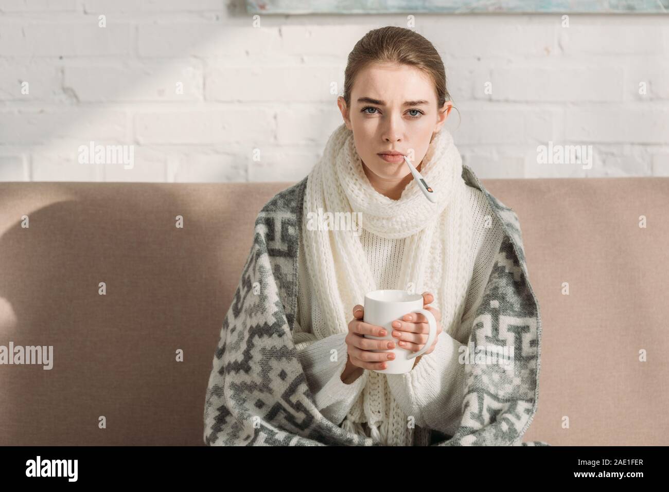 sick girl looking at camera while measuring temperature and holding cup of warming drink Stock Photo