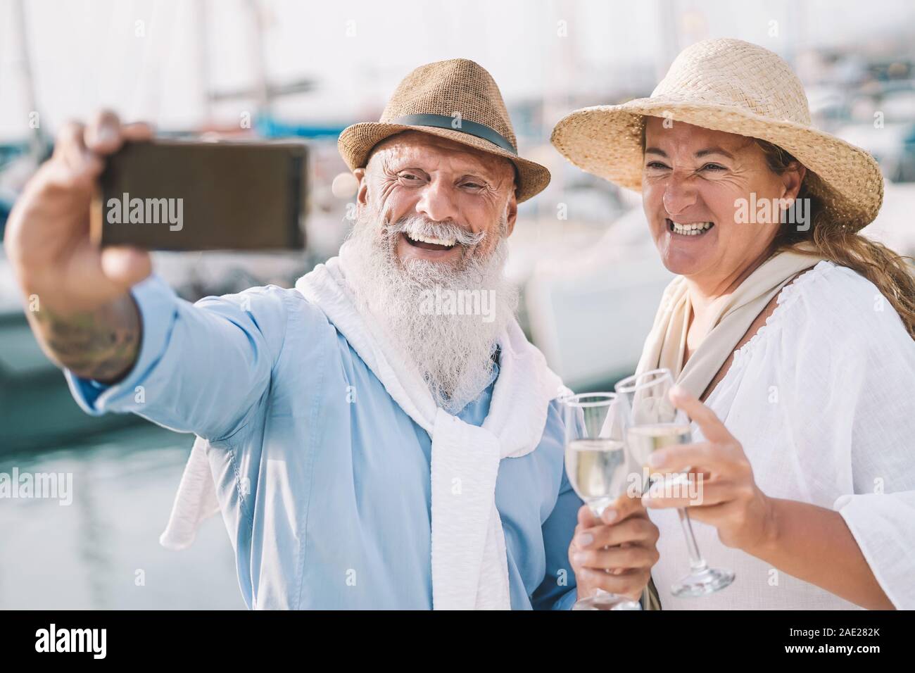 Seniors couple taking selfie with mobile smartphone on sailboat vacation - Happy people having fun celebrate drinking champagne on boat trip Stock Photo