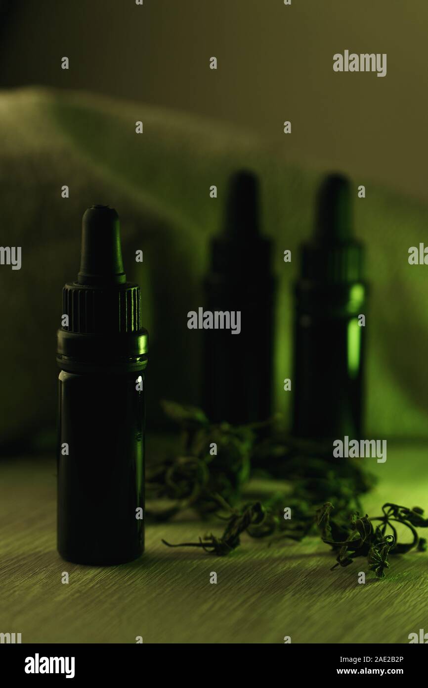 Vertical composition of three 10 ml black glass bottles surrounded with dried herbs on white wooden table backlit with green light Stock Photo