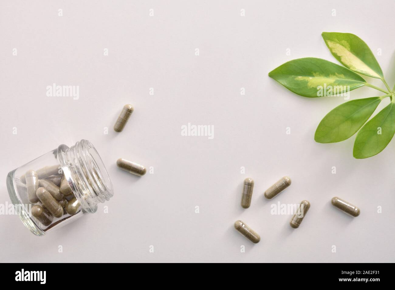 Natural medicine capsules in open glass jar lying on white table and plant leaves. Natural medicine concept. Top view. Horizontal composition. Stock Photo