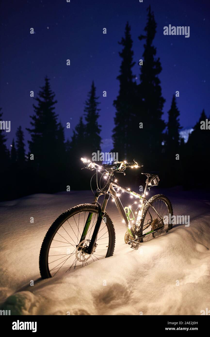 Mountain bike decorated with Christmas lights at snow bank in winter mountains under night sky with stars. Present for New Year. Stock Photo