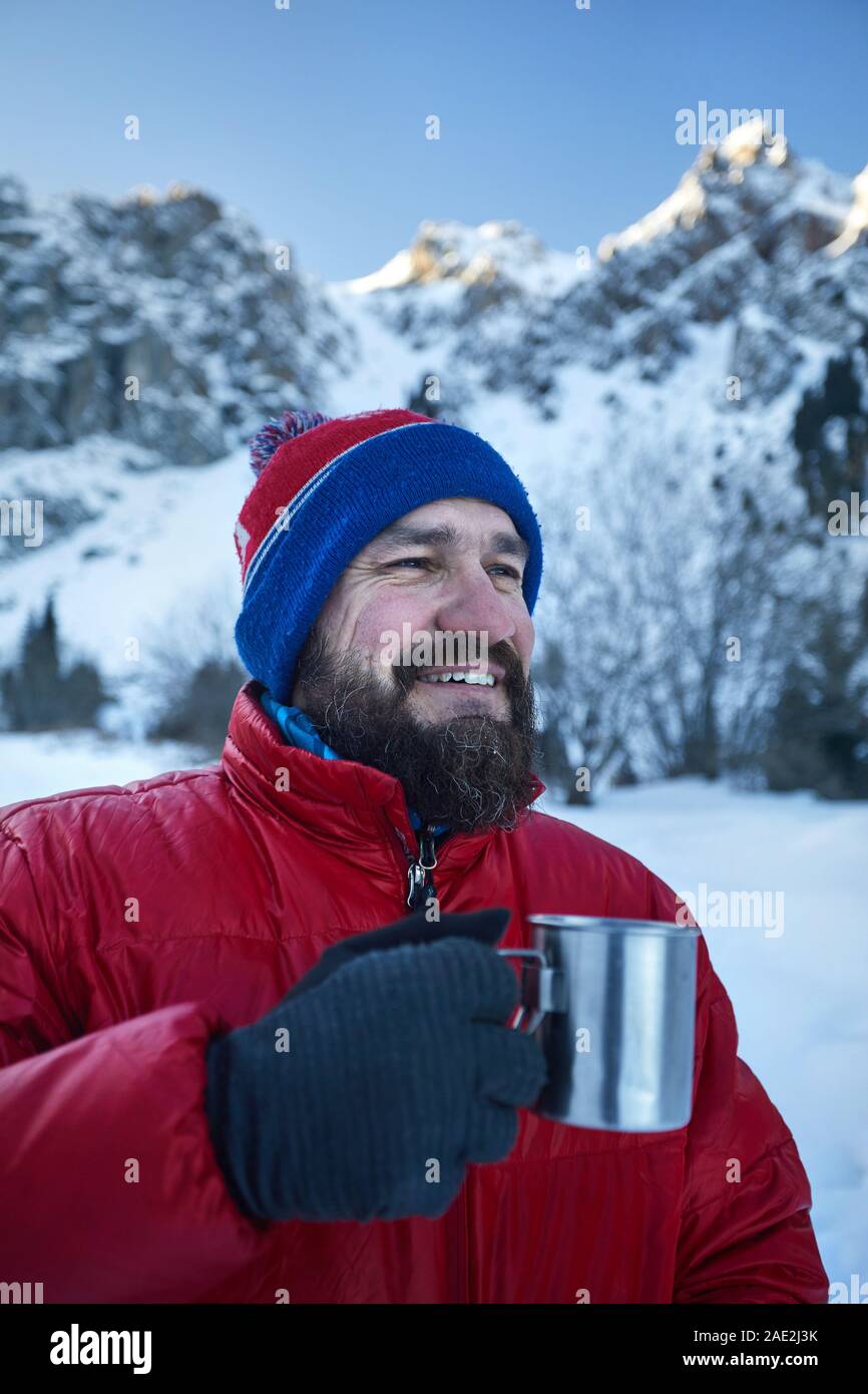 Traveler man with beard is drinking tea or coffee from cup in mountains at winter time. Mountain camping concept. Stock Photo