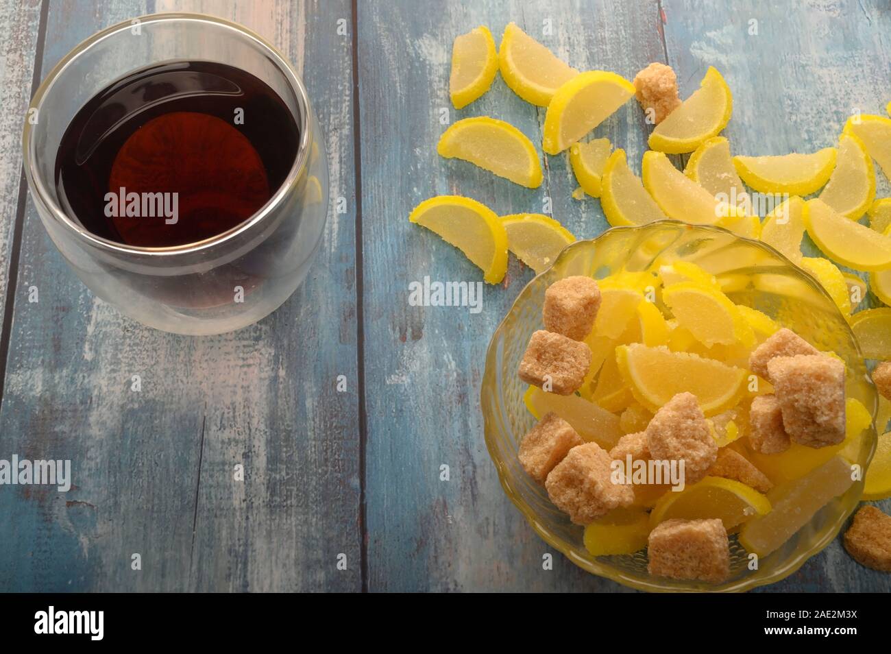 Marmalade lemon slices, pieces of brown sugar and a glass of black tea on a wooden background. Sweet dessert. Close up Stock Photo