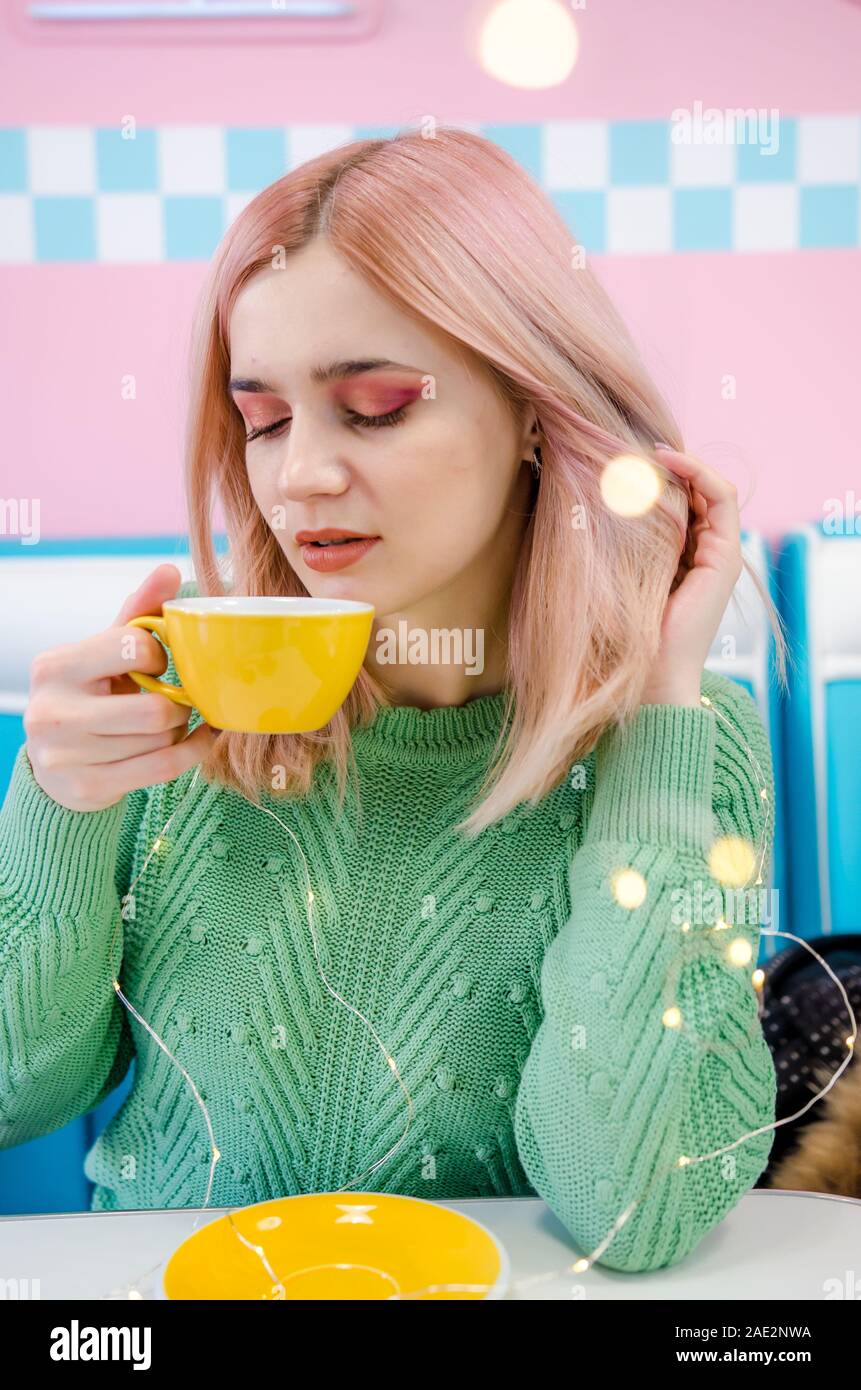 Pretty girl drinking tea in cafe. Portrait of a lovely girl in green sweater with bokeh lights. Pink and blue background Stock Photo