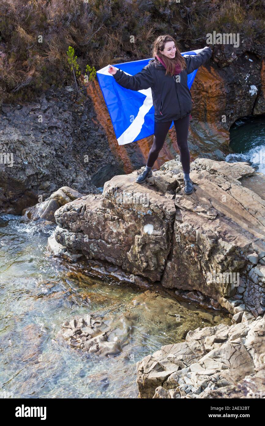 Young woman standing on rocks holding Scottish flag at Fairy Pools, river Brittle, Isle of Skye, Scotland, UK in March Stock Photo