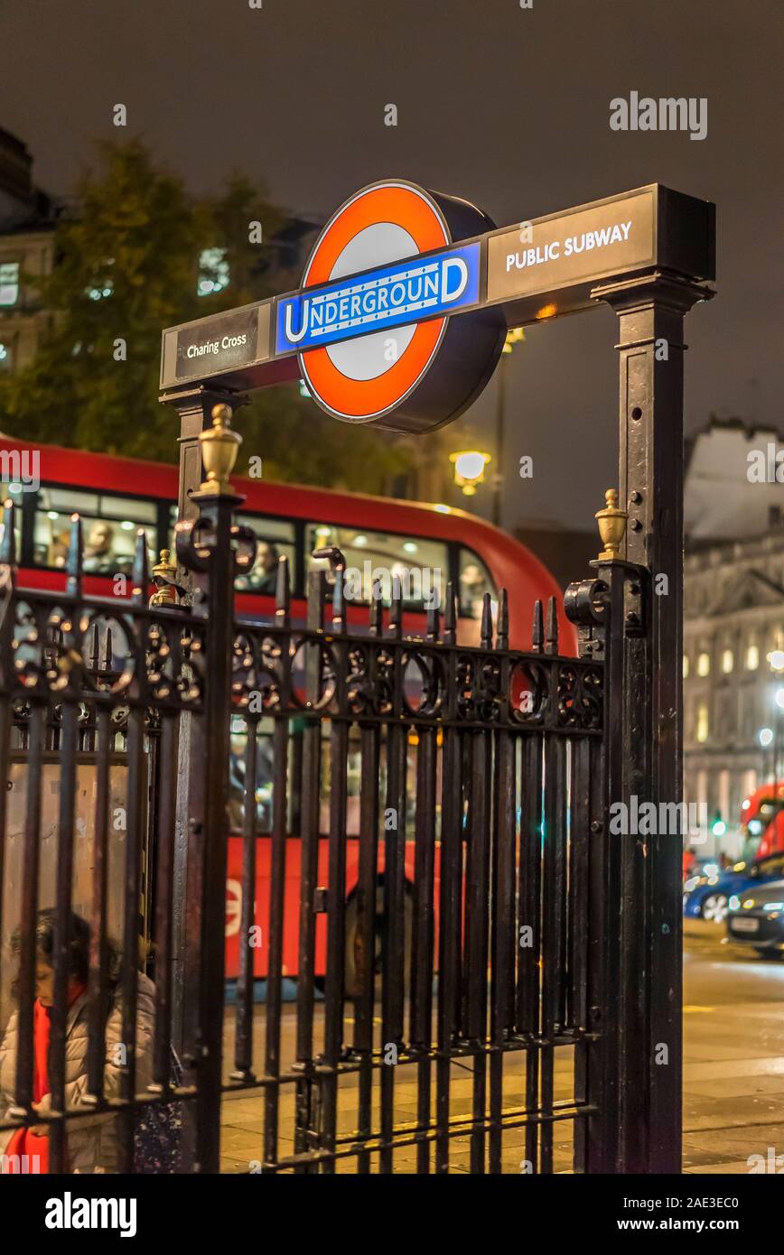 City of London at night. London Underground sign, railings at entrance to Charing Cross Station in Trafalgar Square, central London UK, lit up by lamp. Stock Photo