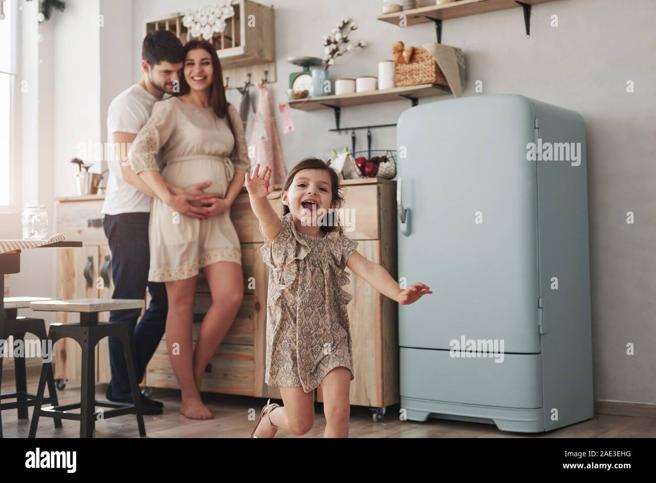 Cheerful people. Playful female child have fun by running in the kitchen at daytime of front of her mother and father Stock Photo