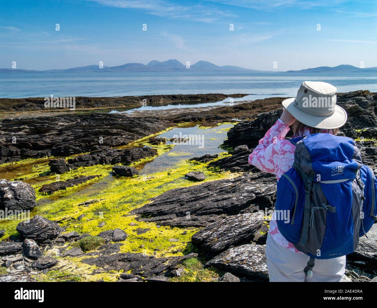Woman holidaymaker looking through binoculars from Colonsay across the sea to the Paps of Jura, Inner Hebrides, Scotland, UK Stock Photo
