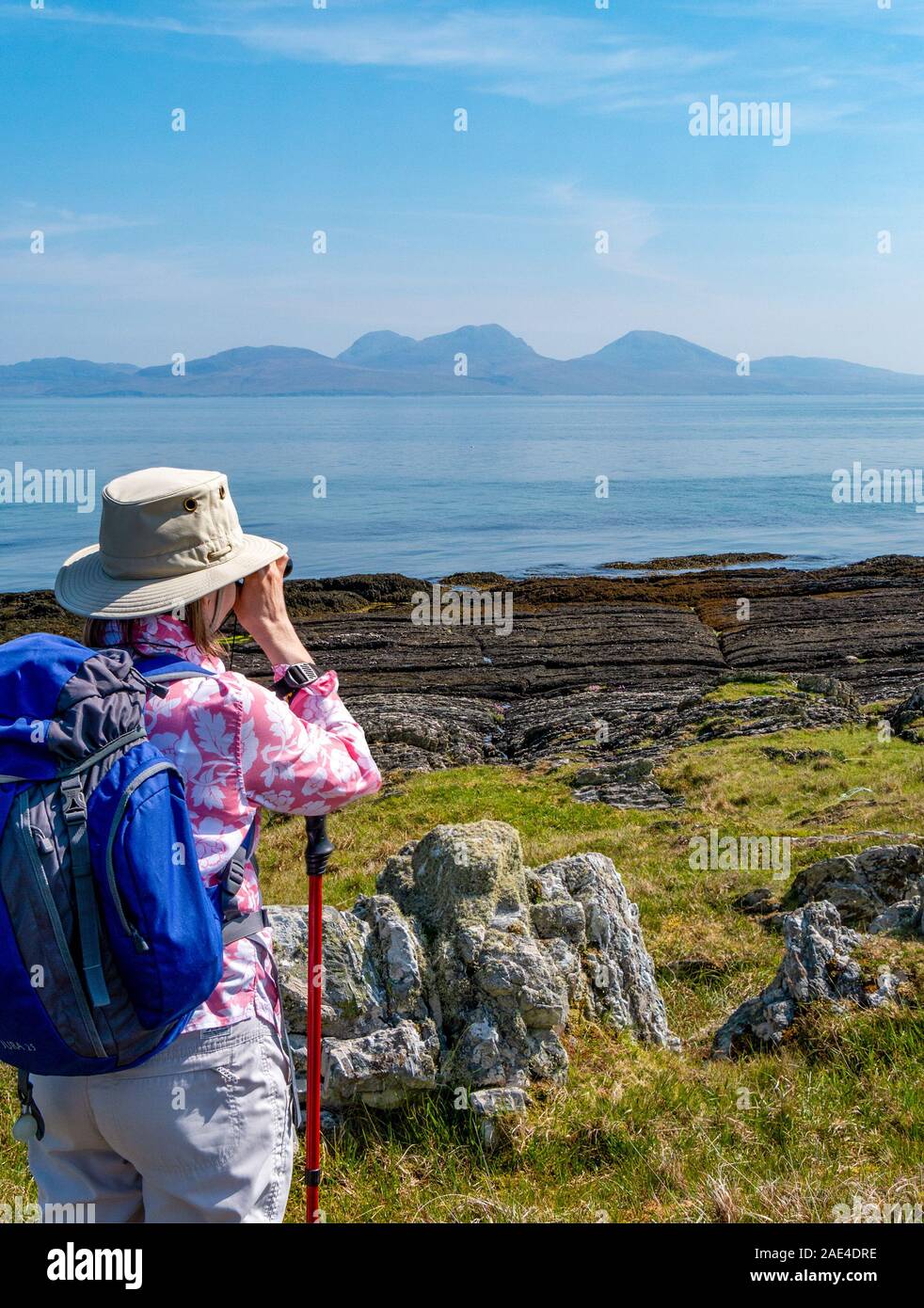 Woman holidaymaker looking through binoculars from Colonsay across the sea to the Paps of Jura, Inner Hebrides, Scotland, UK Stock Photo