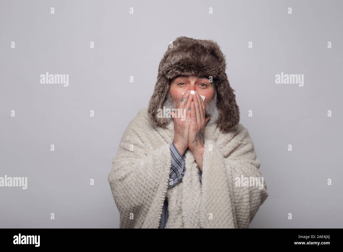 mature man covering himself with a blanket and hat blowing his nose