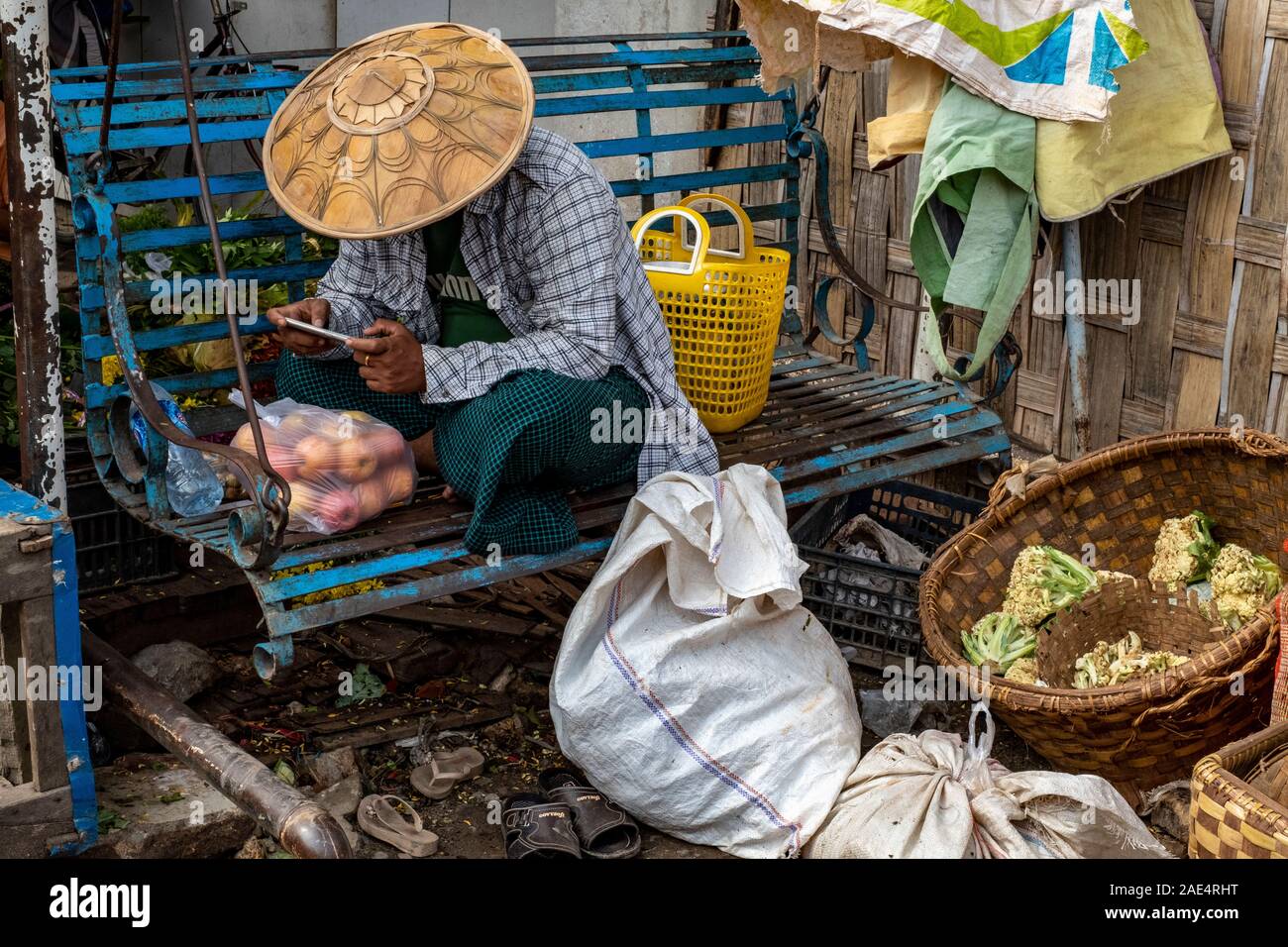 A man in a conical rice hat or coolie hat relaxes on a blue bench with a cell phone after buying apples in the railroad market of Mandalay, Myanmar Stock Photo