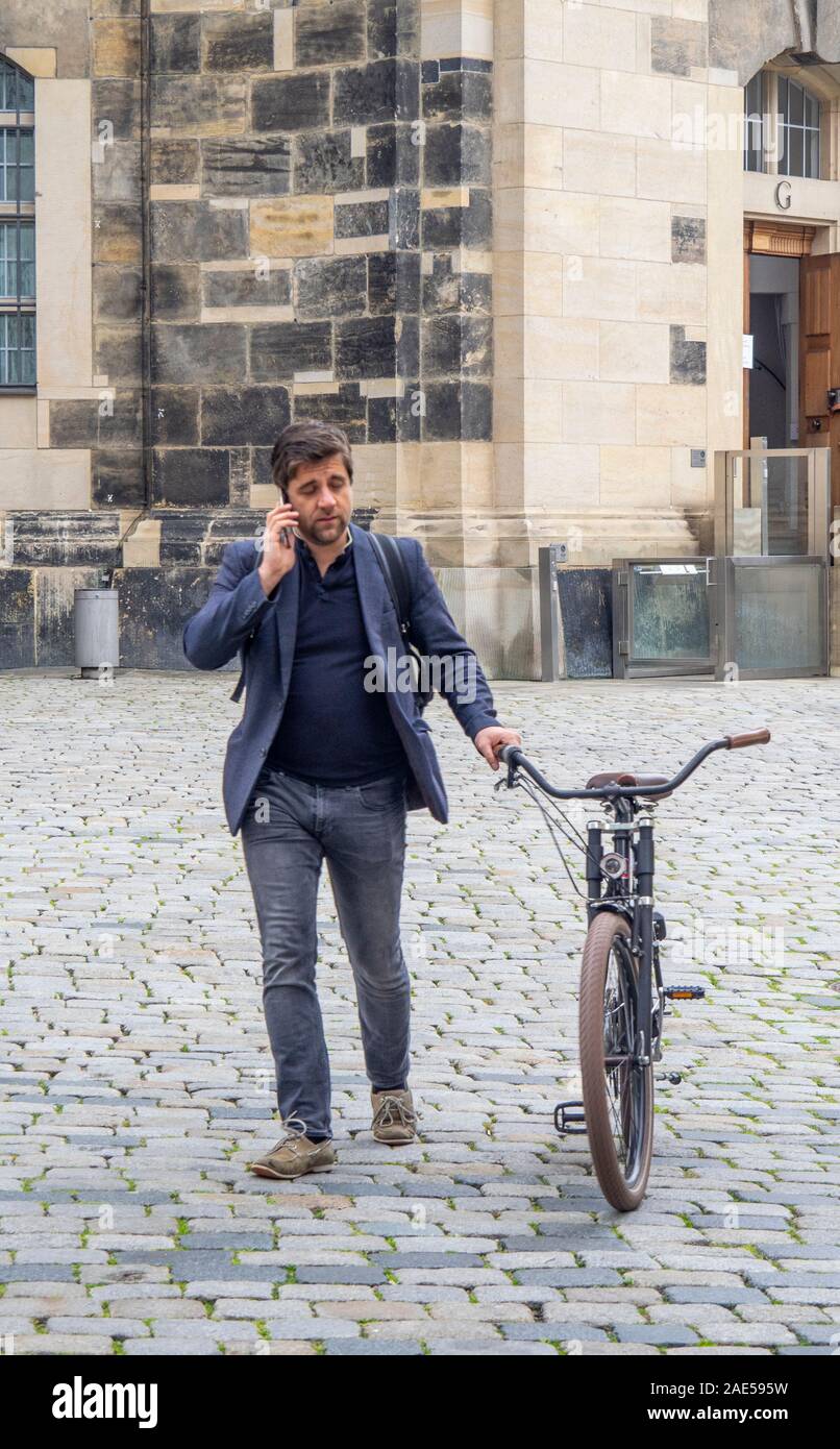 Man talking on mobile telephone and pushing a bicycle across cobblestone Platz Neumarkt Newmarket Altstadt Dresden Saxony Germany. Stock Photo