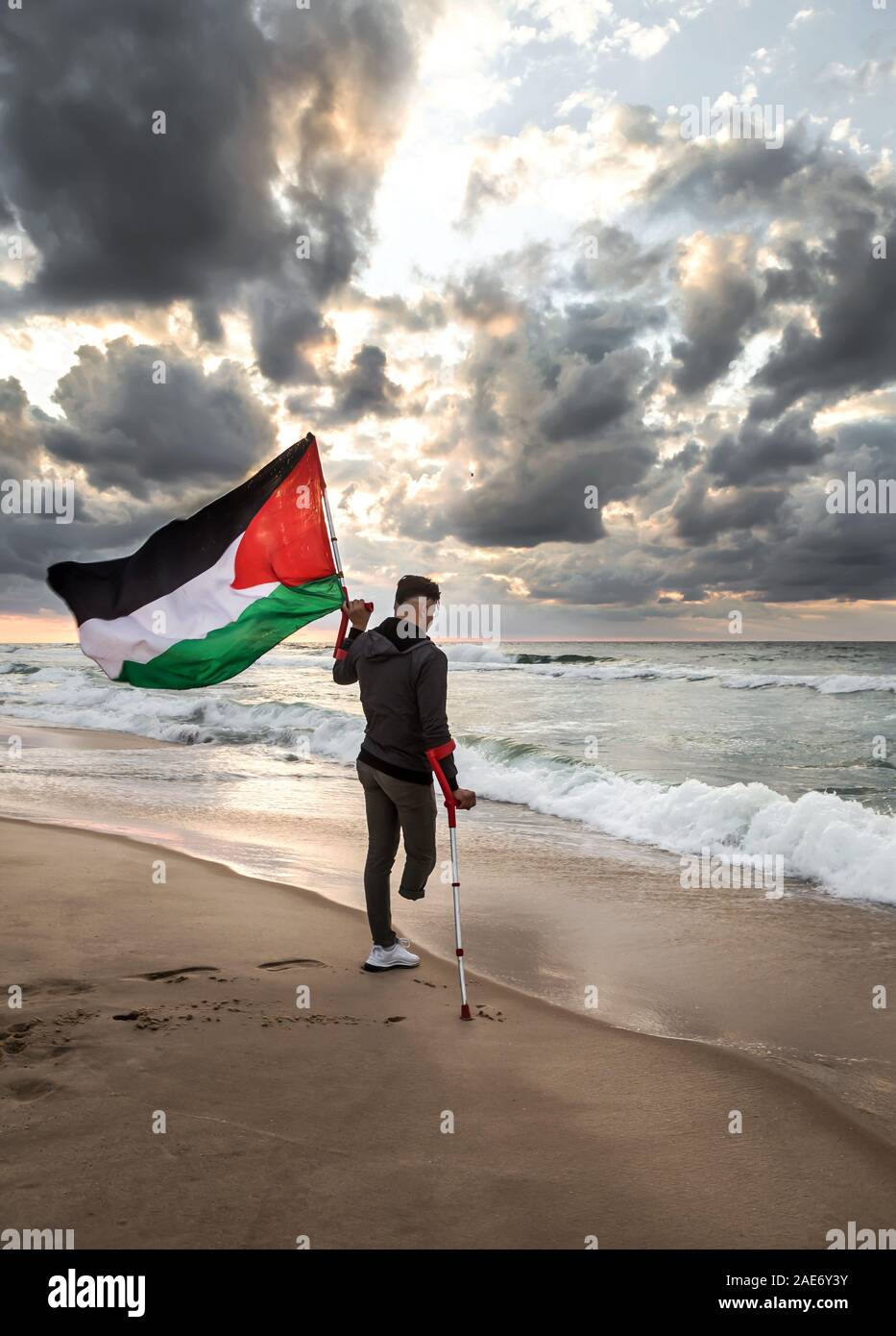 Photo of Palestinian youth with amputated leg, holding up Palestine flag in front of the beach. Stock Photo